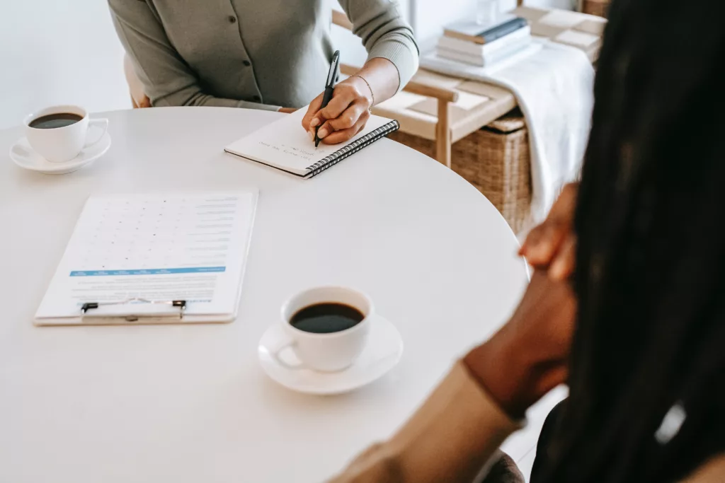 Individuals sitting at a table with a notebook and coffee. Psychological Assessment in New York City with an NYC therapist can help you get the answers you need.